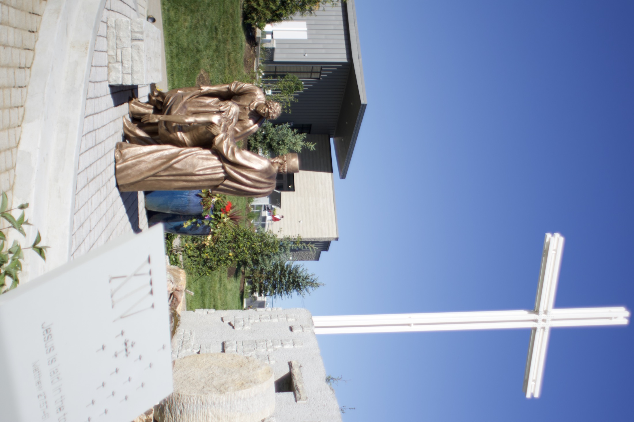 A cross rising against the sky beside a station marker inscribed with raised braille, the Welcome Center beyond.