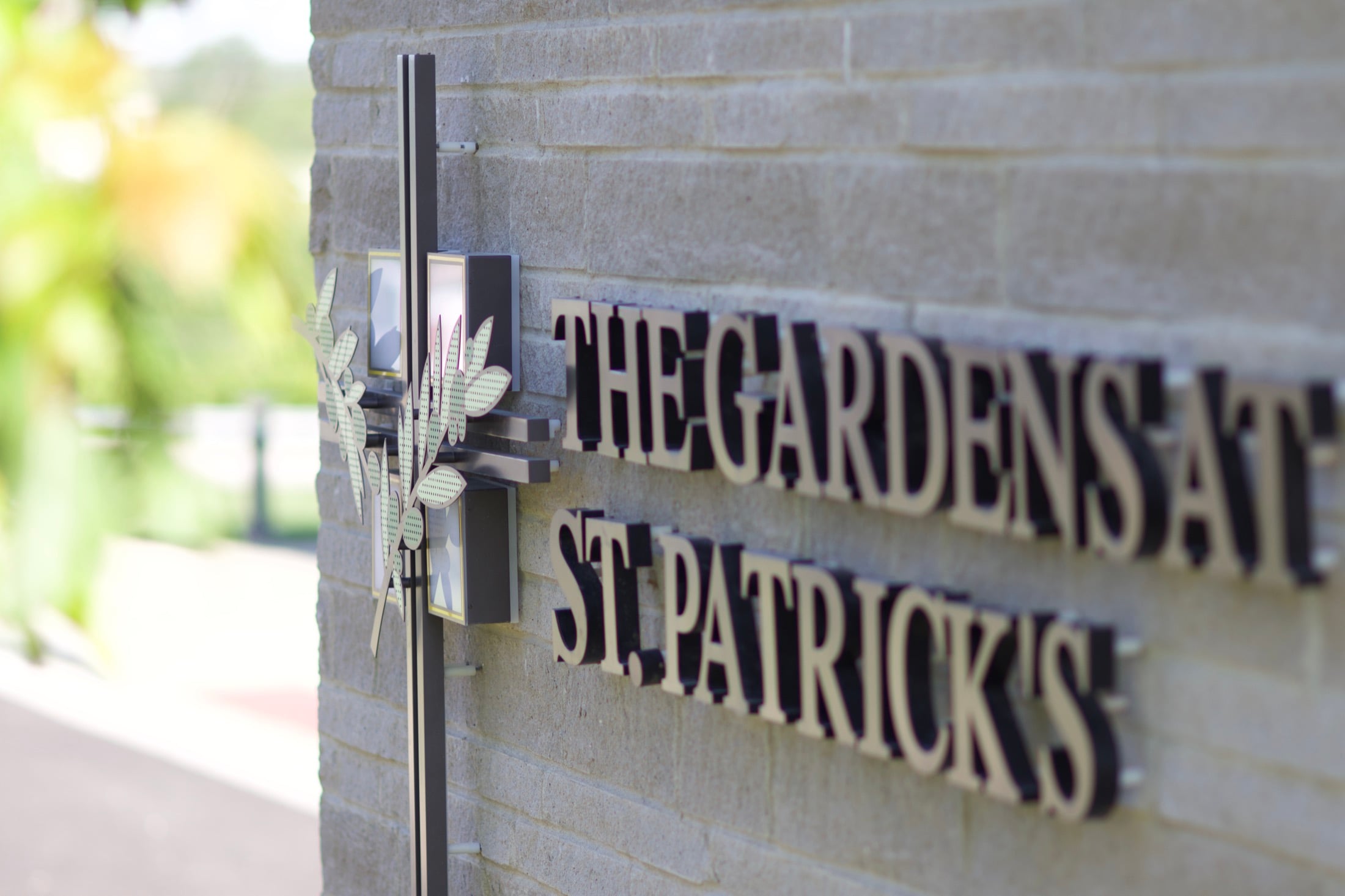 A carved stone sign reading The Gardens at St. Patrick's, mounted beside an olive branch bracket at the entrance.