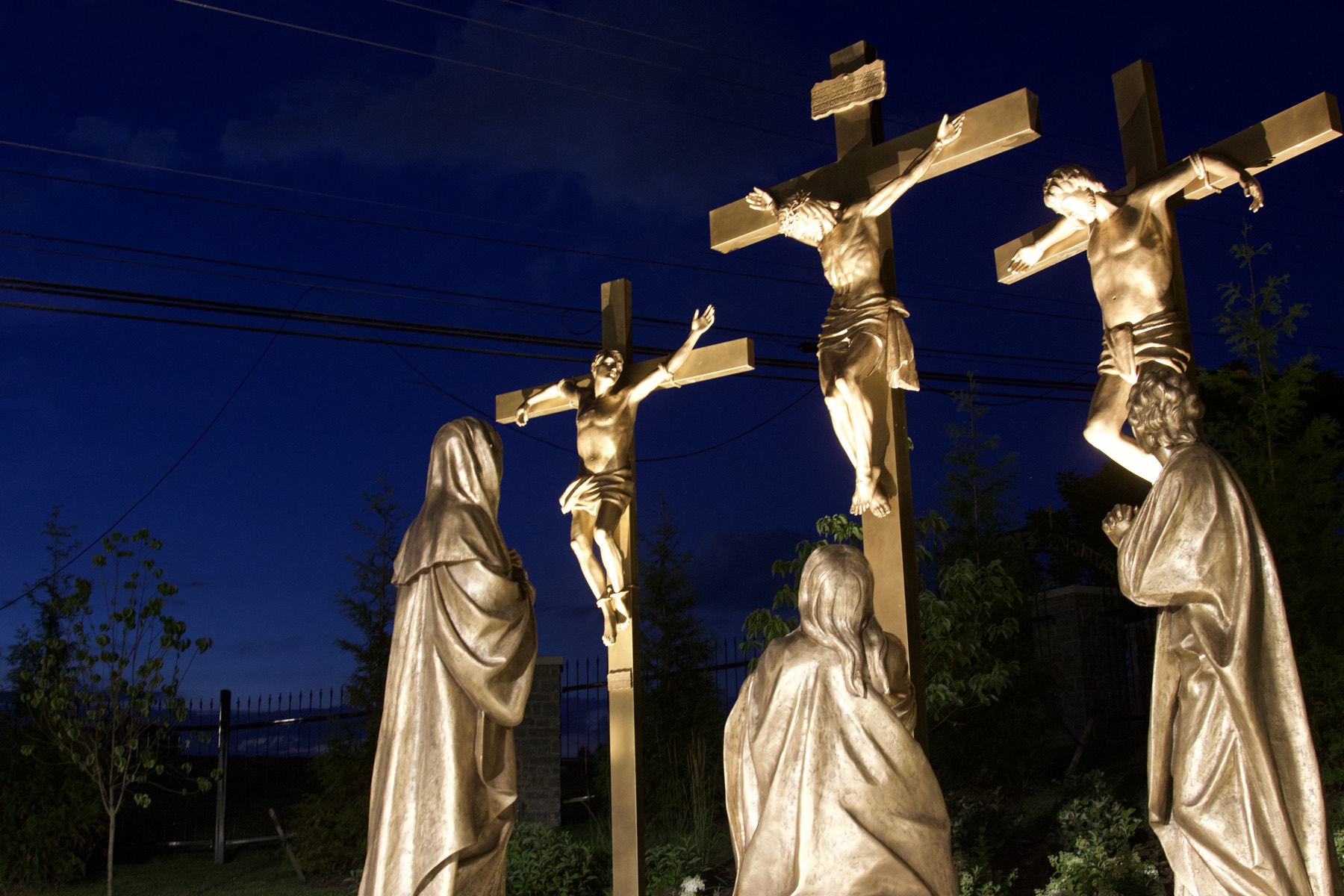 Three bronze crucifixion crosses against a dusk sky, mourning figures gathered below.