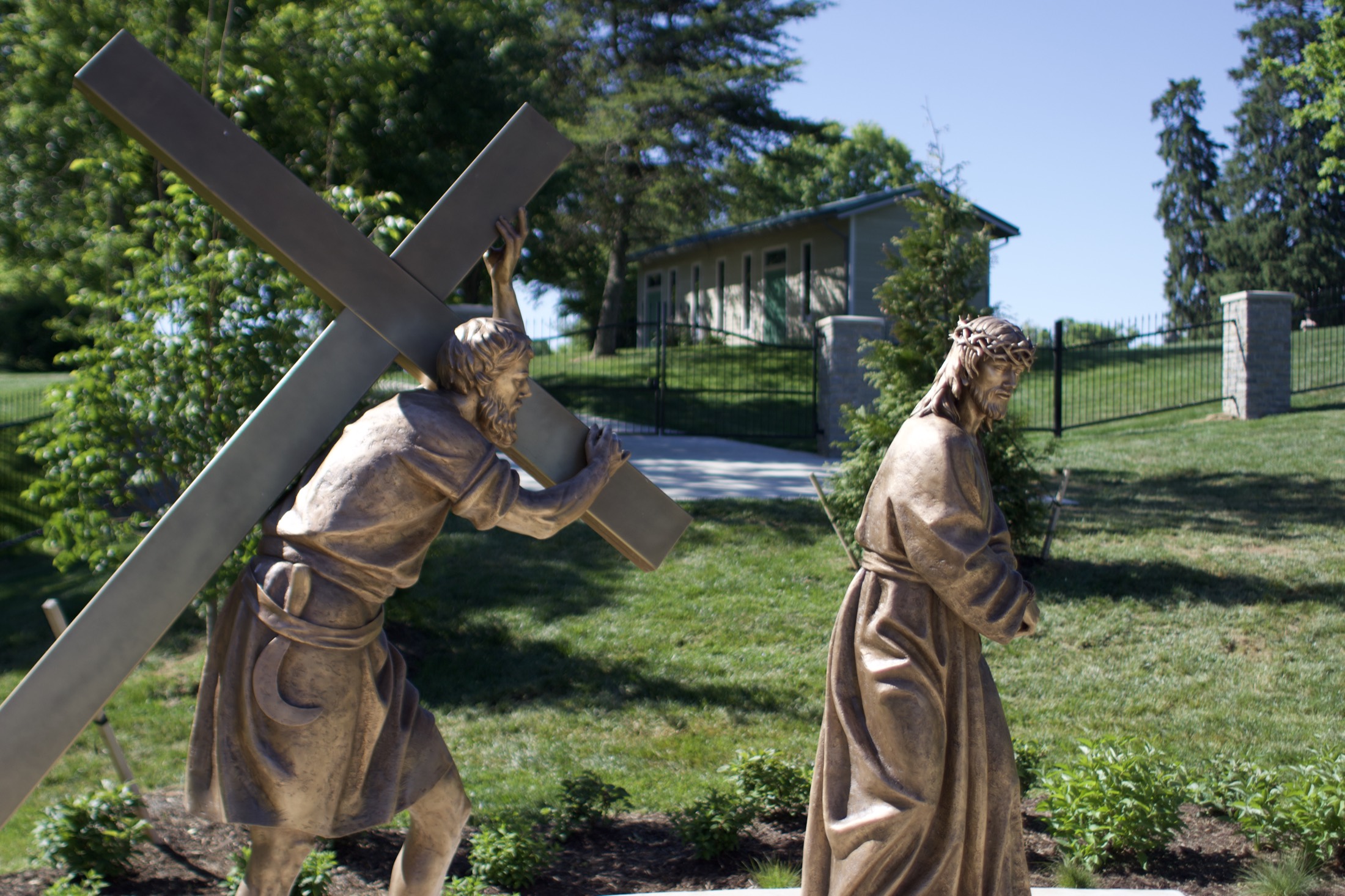 A bronze figure of Christ taking up the cross, the illuminated monumental cross glowing beyond at dusk.