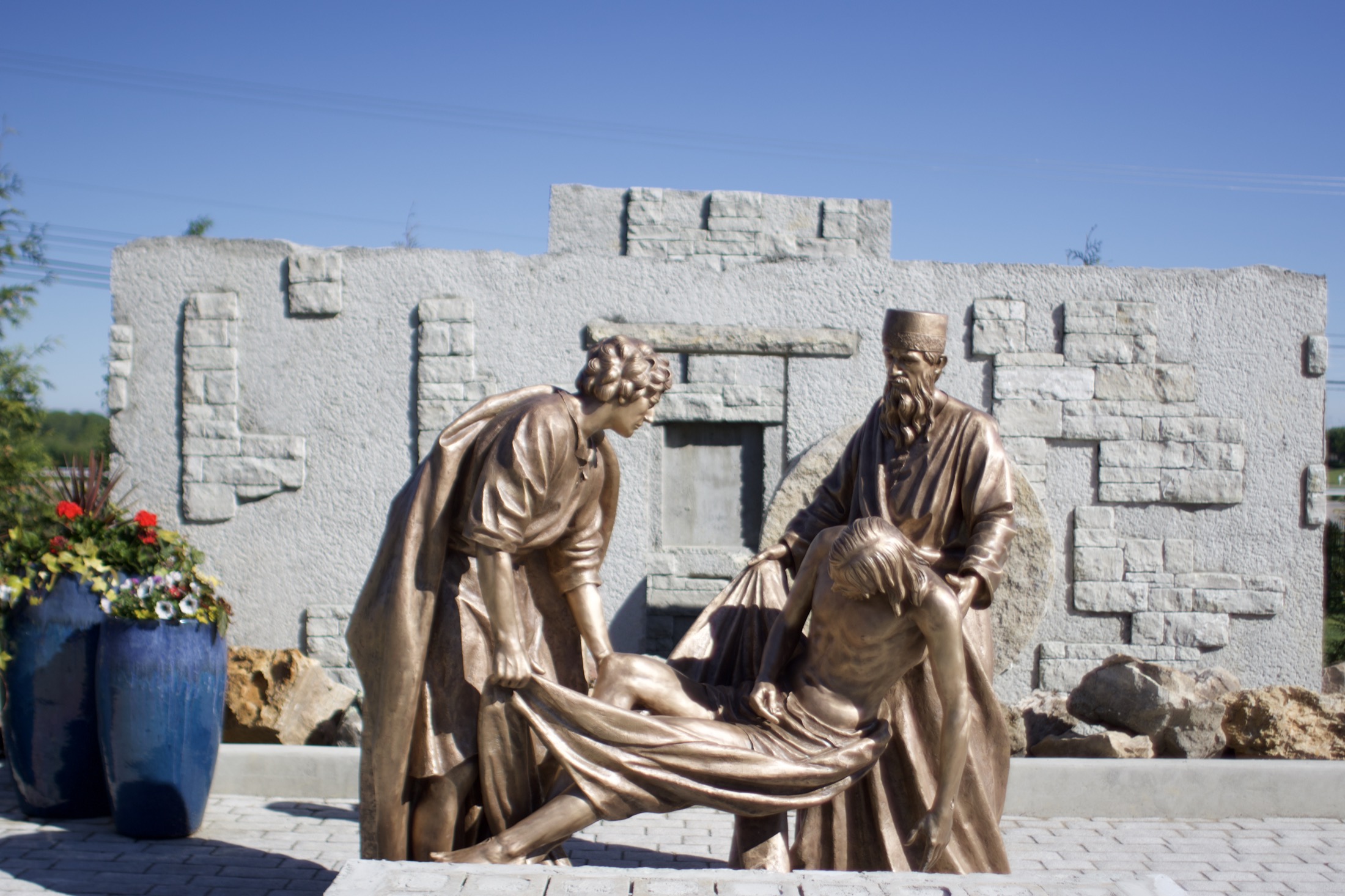 Two bronze figures lowering the body of Christ, beside blue planters of flowers.