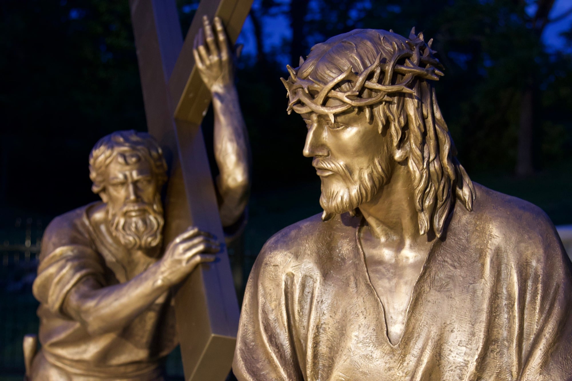 Bronze figures of Christ and Simon of Cyrene together at the cross, lit against a deep blue evening sky.