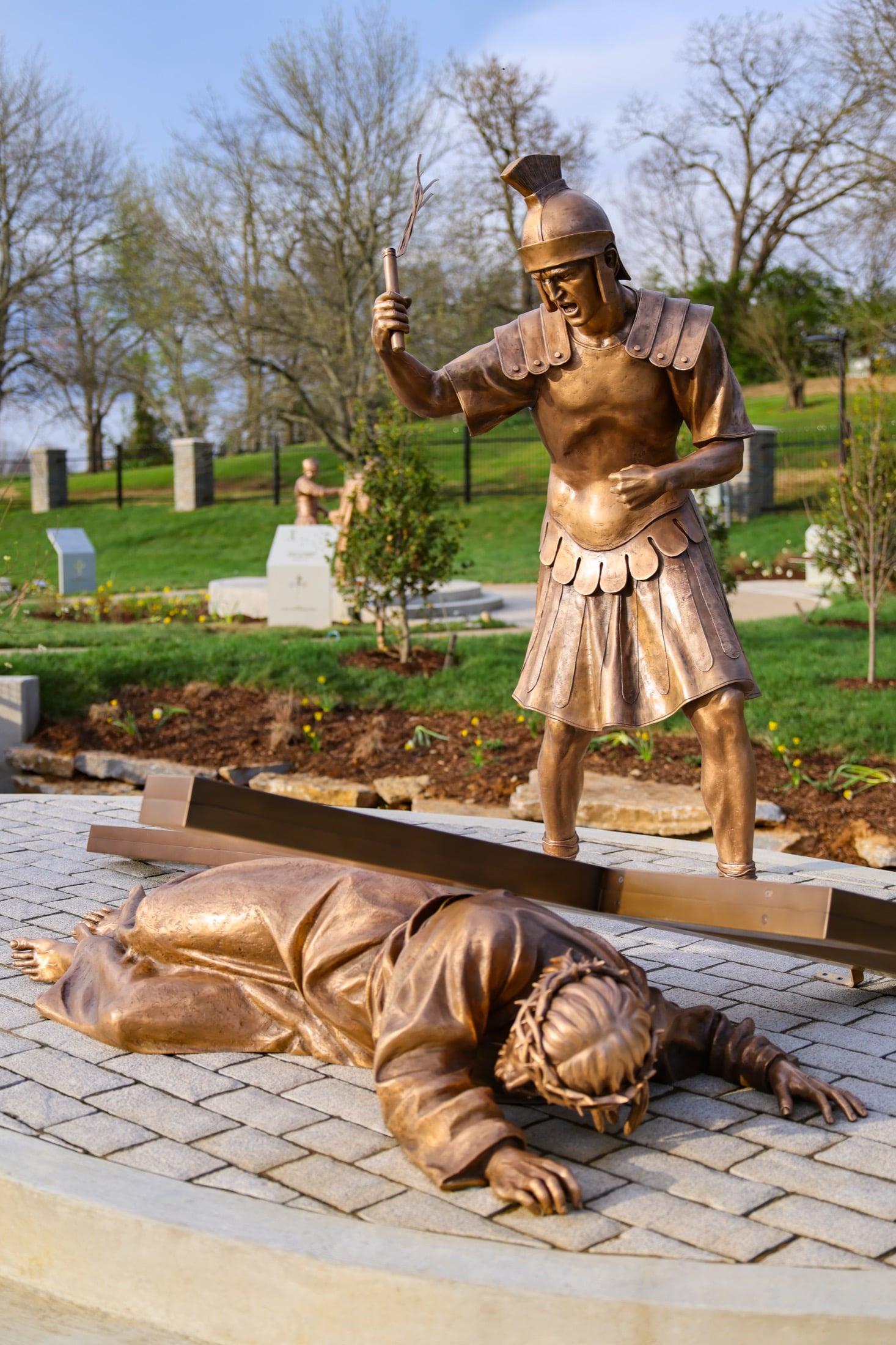 A bronze Roman soldier standing over Christ, who has fallen beneath the weight of the cross.