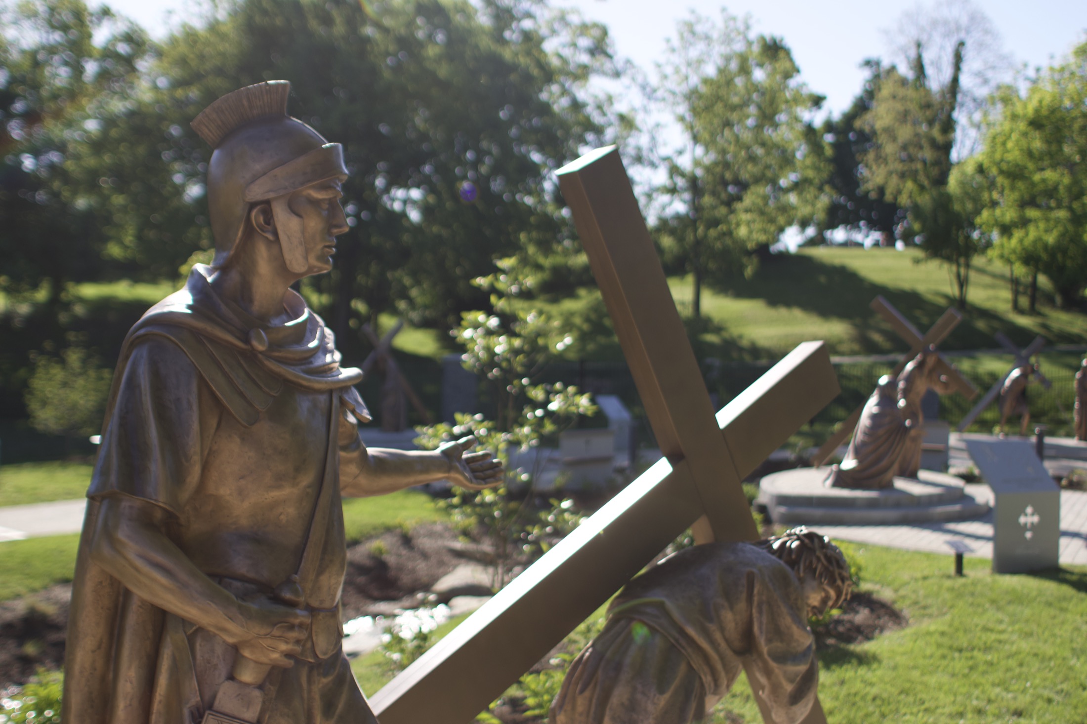 A bronze Roman soldier and Christ carrying the cross, set in the open garden in daylight.