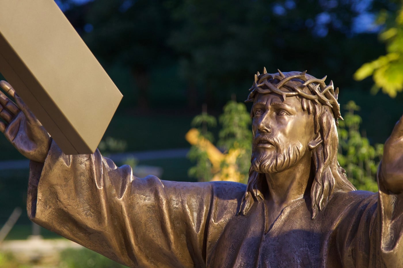 A bronze figure of Christ carrying the cross, catching warm light against the evening garden.