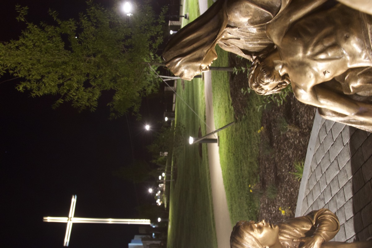 A bronze Pietà beside the lit garden path at night, the monumental cross glowing beyond.