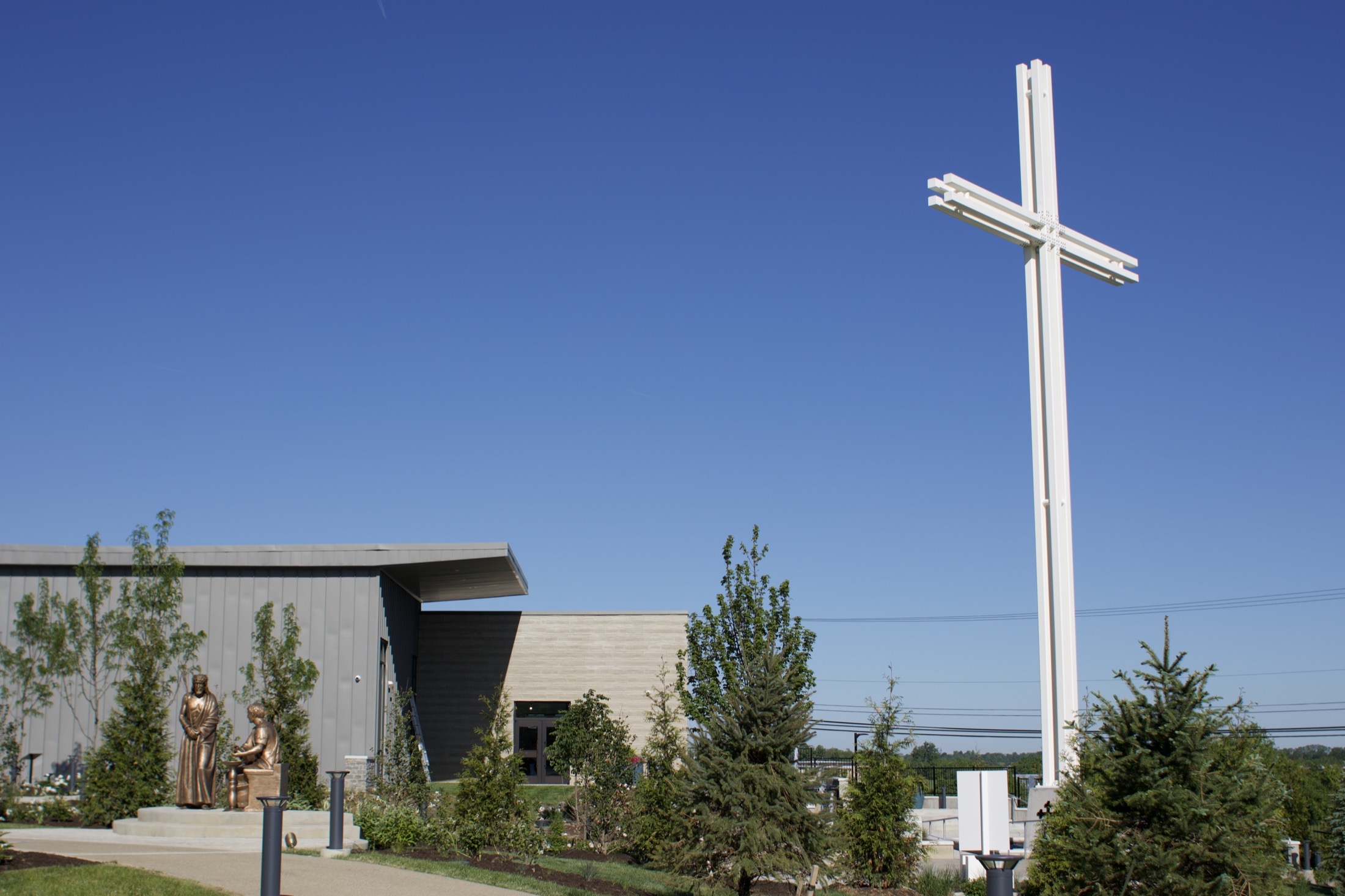 The Gardens at St. Patrick's Welcome Center building beside the monumental cross, rising against a clear blue sky.